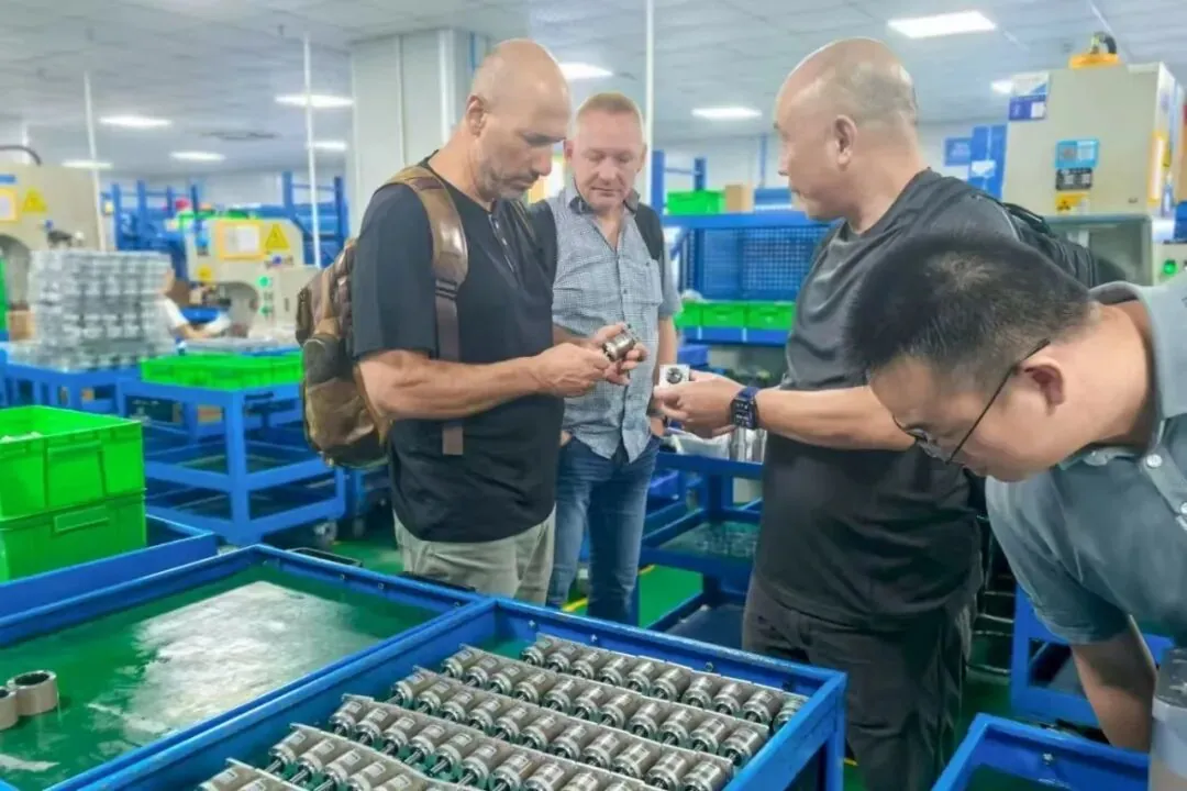 Four men inspect small mechanical parts on the floor of a brightly lit manufacturing facility.