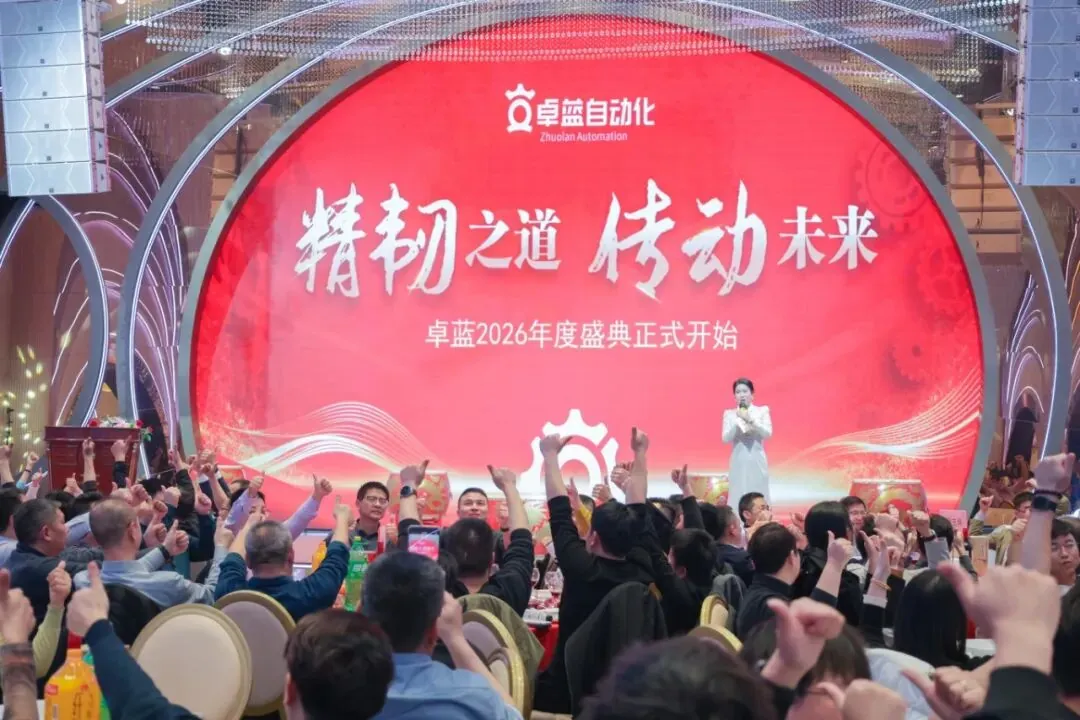 An indoor event with a large red screen displaying white text, featuring a woman speaking on stage to an audience with many hands raised in a thumbs-up gesture.