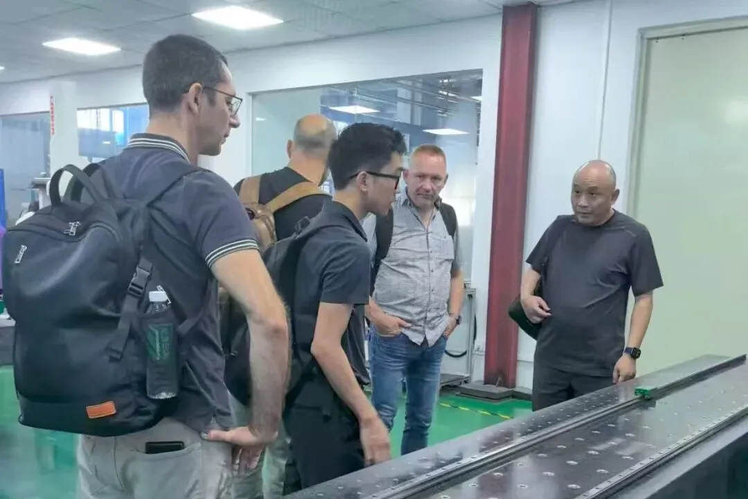 Four men stand in a manufacturing facility, with one man pointing to a long, flat surface.