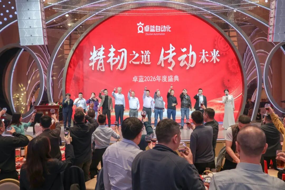 An indoor event with a group on stage raising glasses, facing an audience who are also raising glasses for a toast beneath a large red screen.