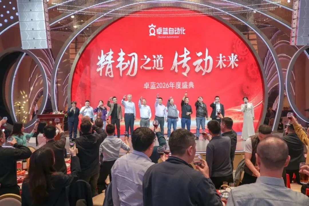 An indoor event with a group on stage raising glasses, facing an audience who are also raising glasses for a toast beneath a large red screen.