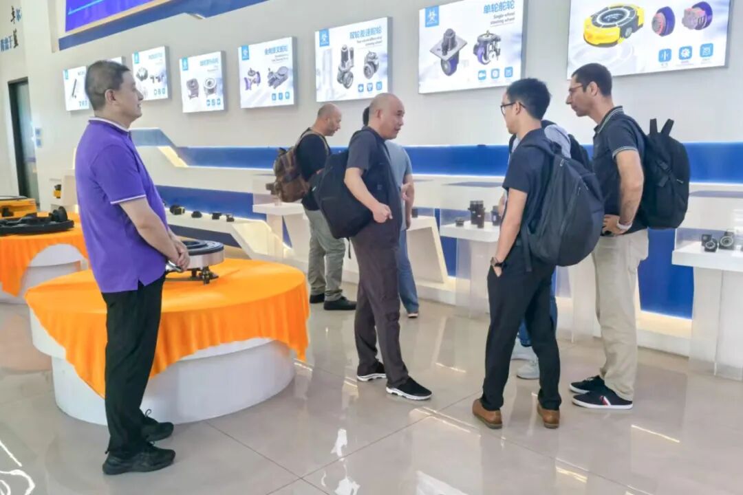 A man in a purple shirt stands near an orange-draped display table in a showroom with several people viewing exhibits.