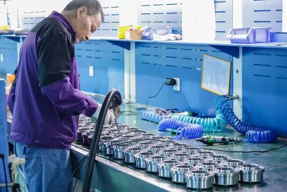 A worker in a purple jacket and blue jeans is working at an assembly station with many silver metal components lined up on the table.