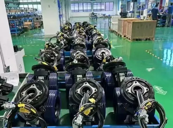 Rows of assembled black mechanical drive units with attached wires are displayed on blue carts on a glossy green factory floor.