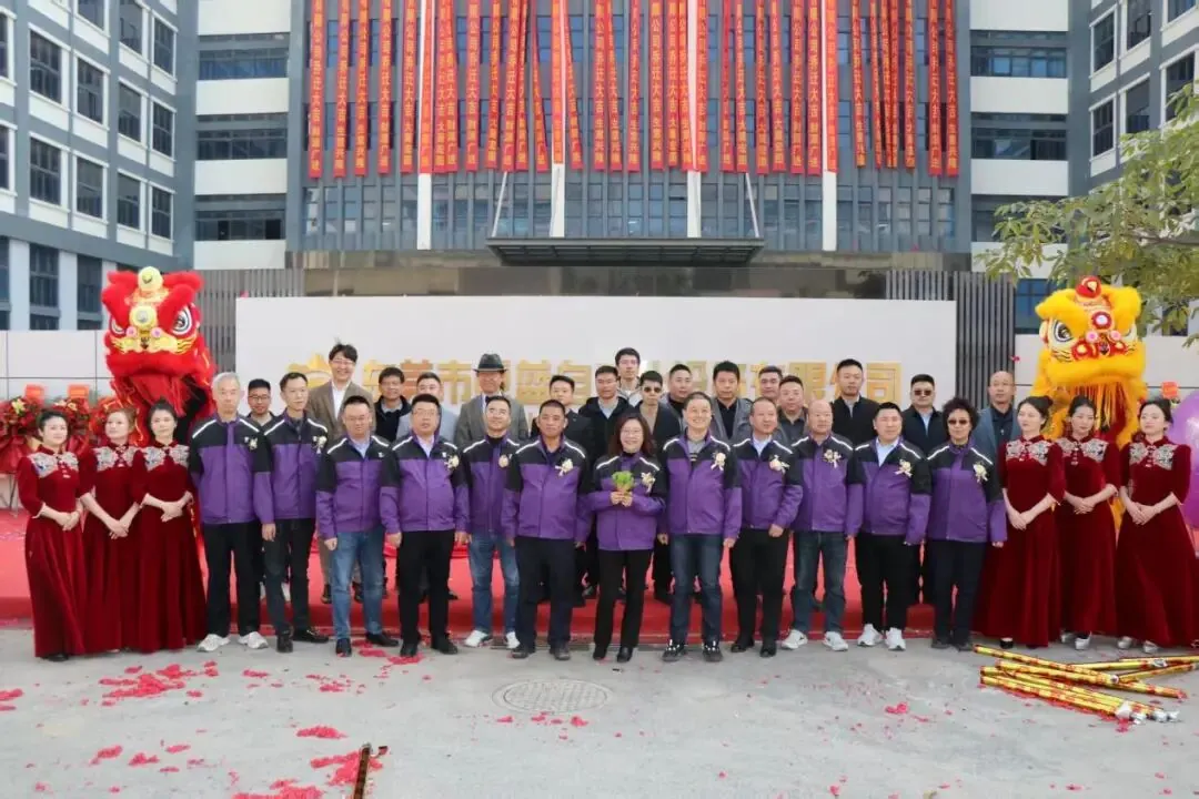 A large group of people wearing matching purple jackets poses outdoors in front of a modern building with red banners.