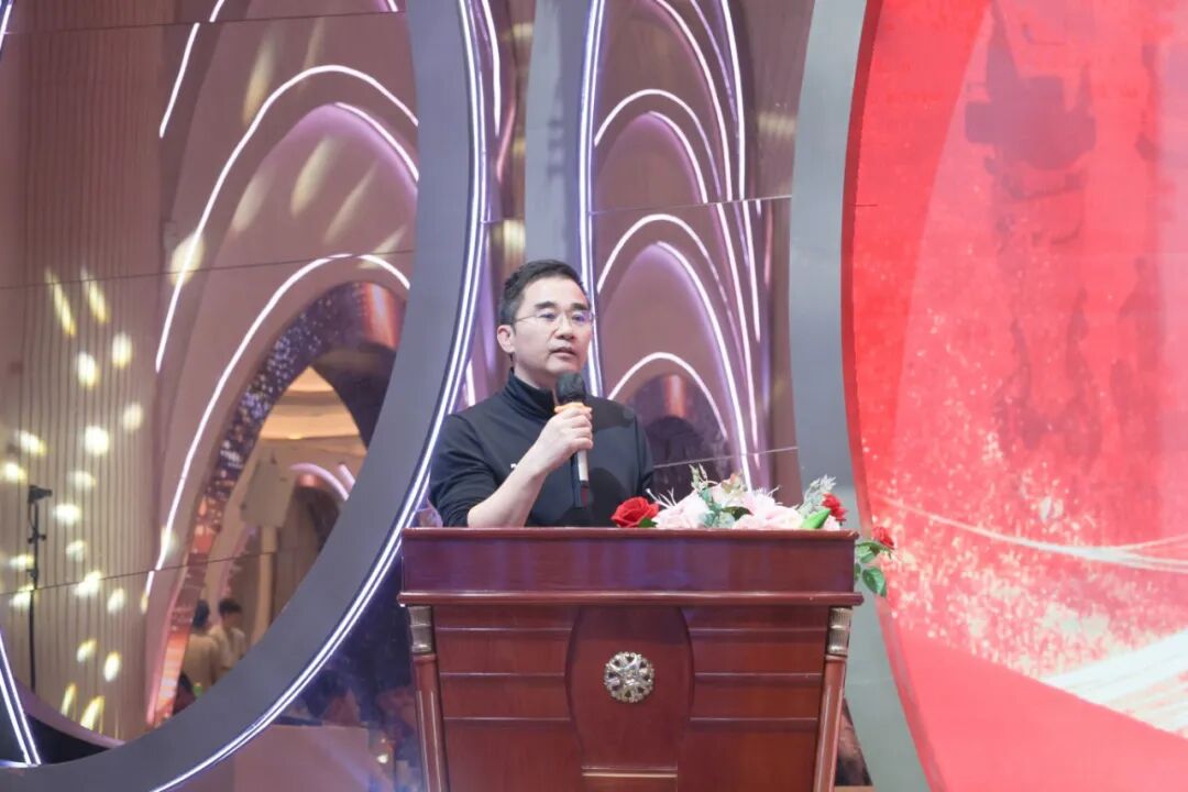 A man wearing glasses and a black shirt speaks into a microphone while standing behind a wooden podium with flowers.