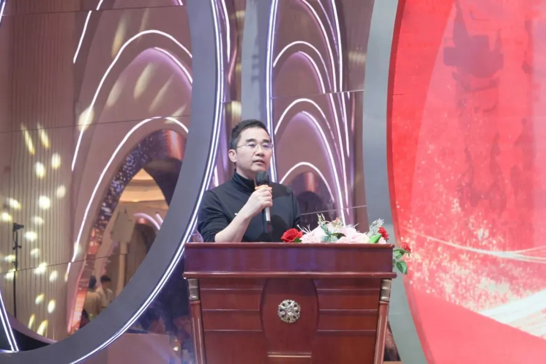 A man wearing glasses and a black shirt speaks into a microphone while standing behind a wooden podium with flowers.