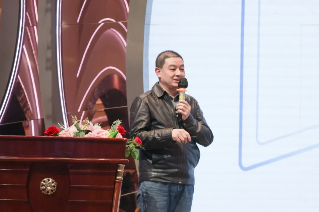 A man in a black leather jacket and jeans stands holding a microphone near a wooden podium with flowers.