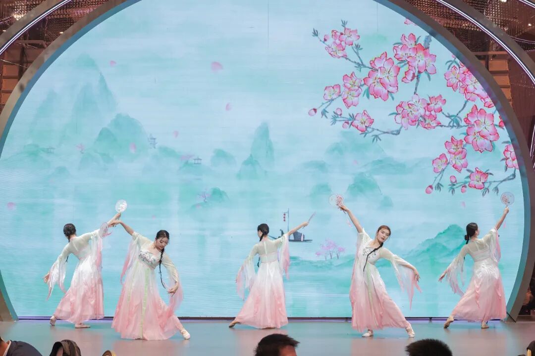 Five dancers in flowing white and pink traditional costumes perform with fans on a stage against a screen displaying a misty landscape with cherry blossoms.