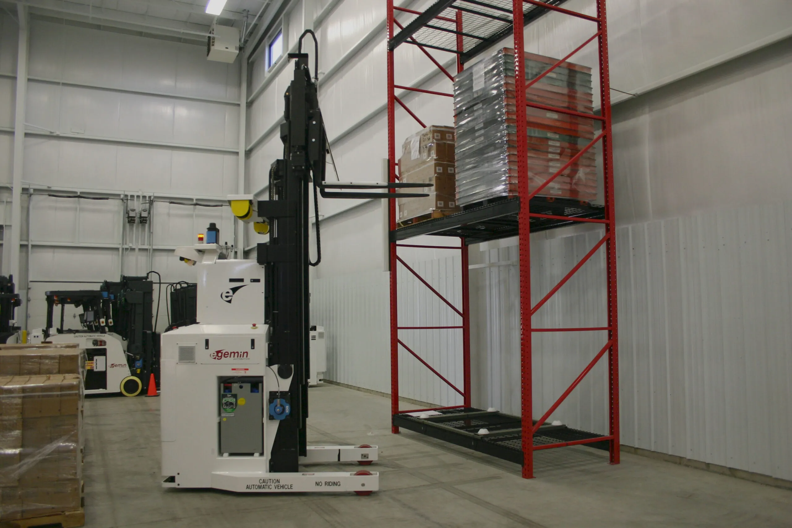 White and black Gemini Automated Stacking Crane positioning forks near a tall red storage rack holding wrapped pallets inside a clean industrial warehouse.