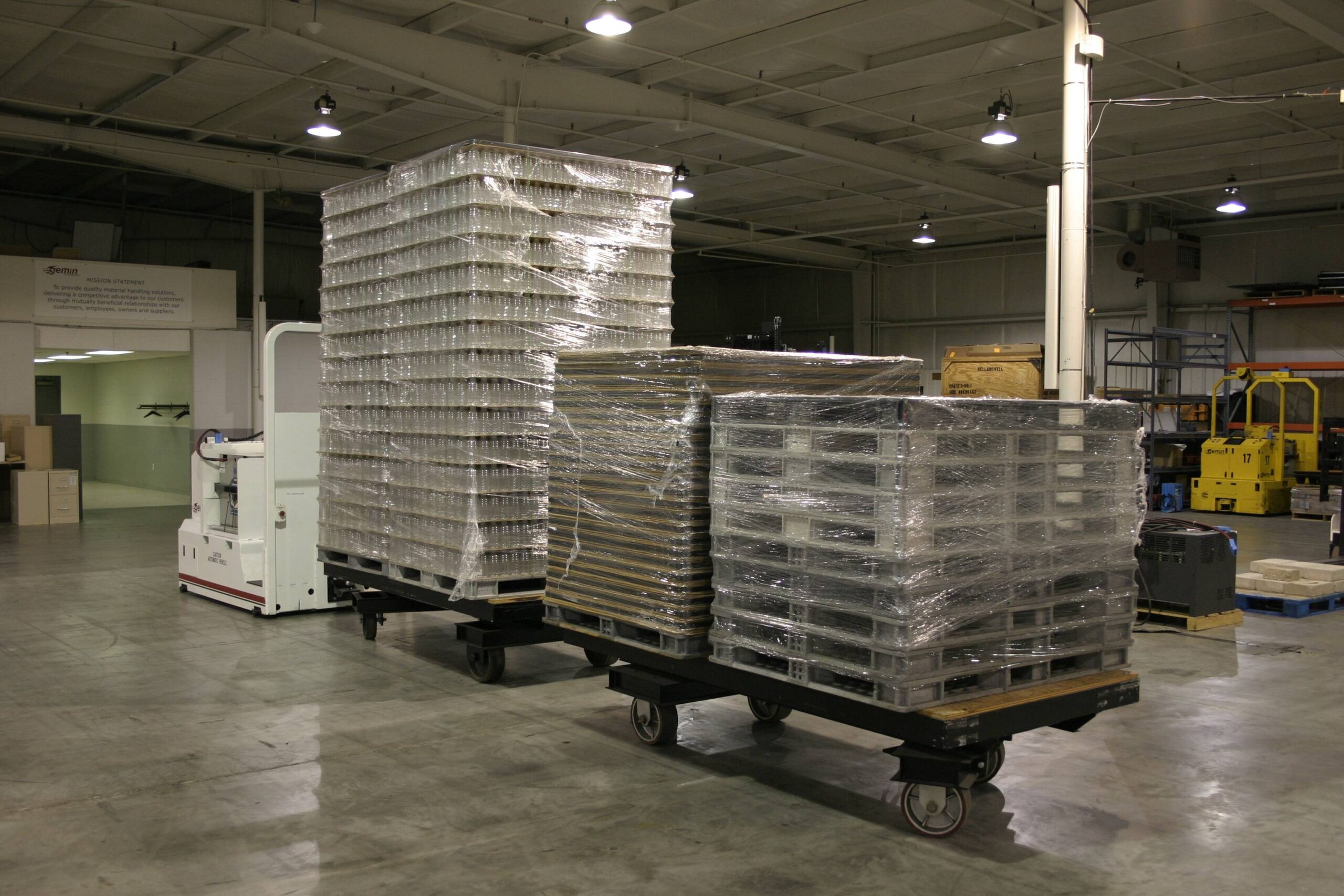 Automated Guided Vehicle towing three carts loaded with tall stacks of plastic containers and wrapped pallets in a brightly lit warehouse with a Gemini sign on the wall.