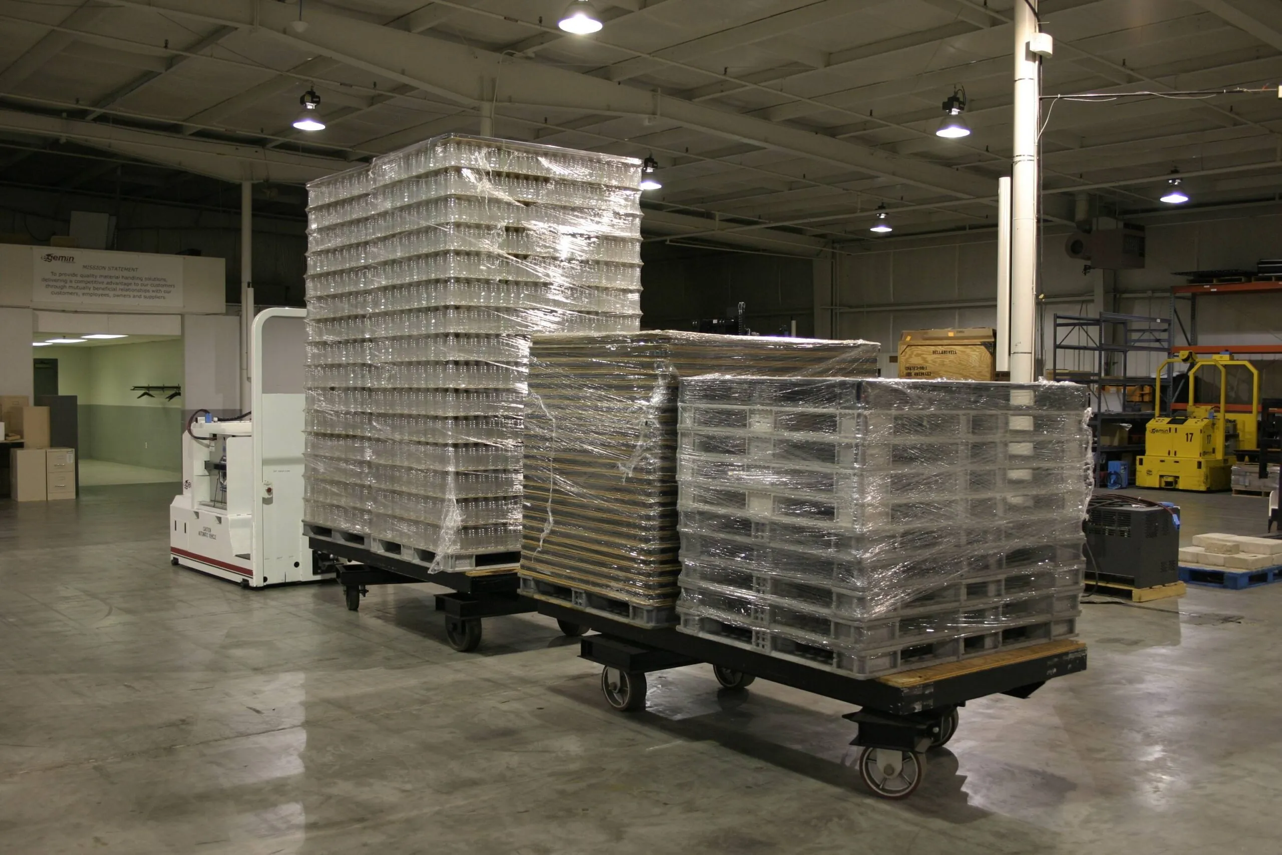Automated Guided Vehicle towing three carts loaded with tall stacks of plastic containers and wrapped pallets in a brightly lit warehouse with a Gemini sign on the wall.