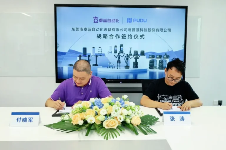 Two men sitting at a white table signing documents in front of a screen displaying industrial robots and automation.