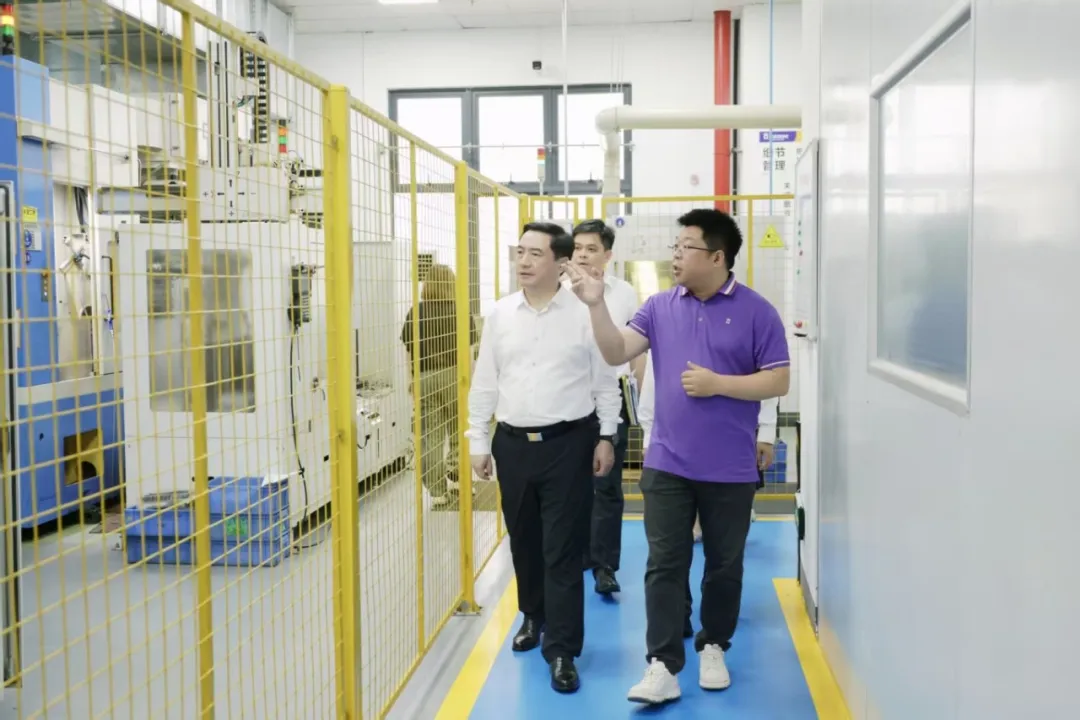 A group of men walk through a modern factory facility while a man in a purple shirt gestures toward industrial machinery.