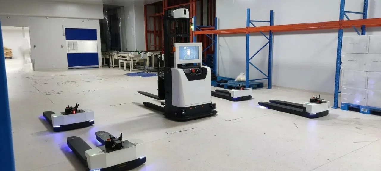 Automated warehouse scene with multiple white and black AGV pallet jack robots and an AGV forklift operating on a clean warehouse floor.