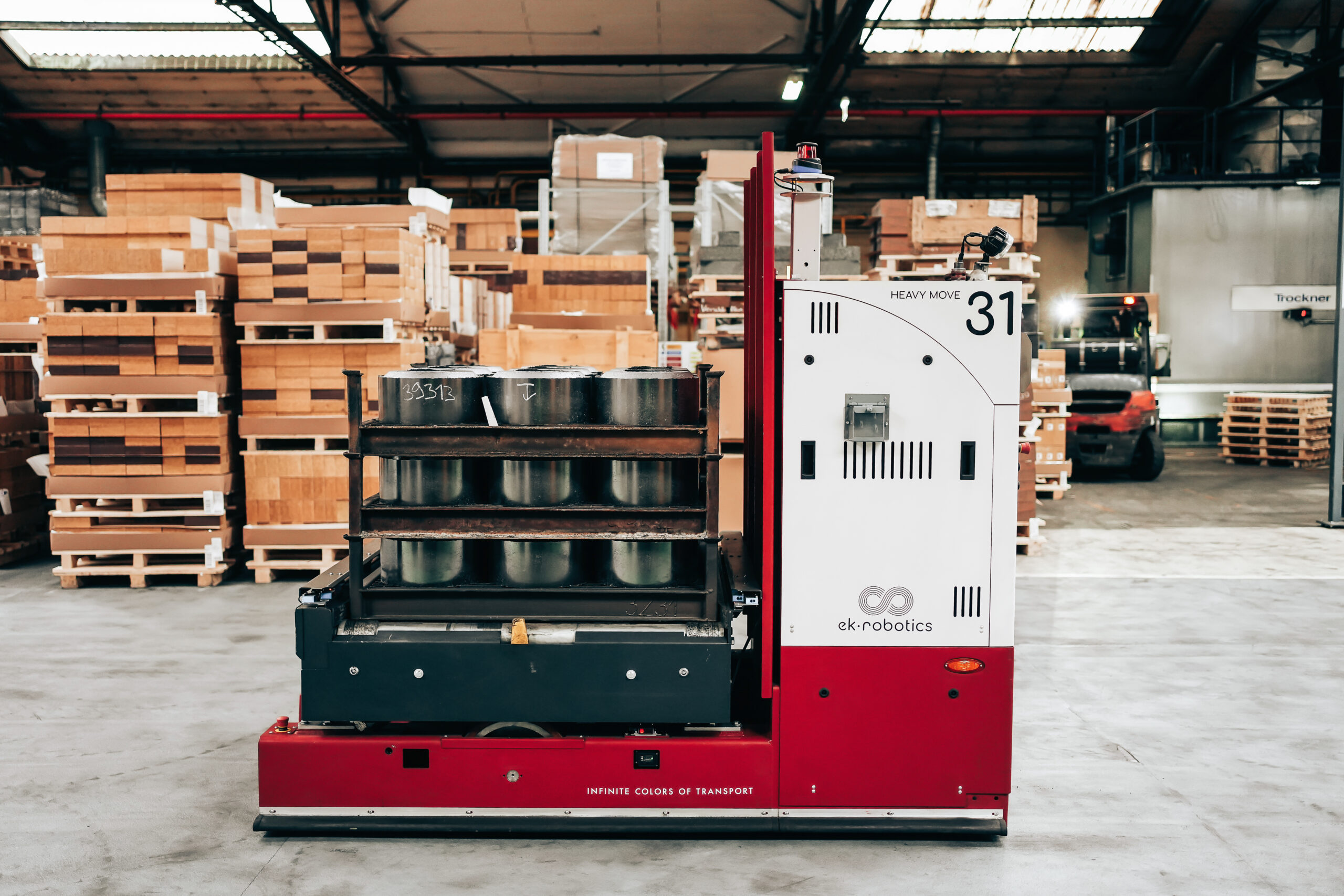 A red and white AGV labeled "HEAVY MOVE 31" carrying metal pots on a rack in a busy warehouse with stacked pallets and a forklift in the background.