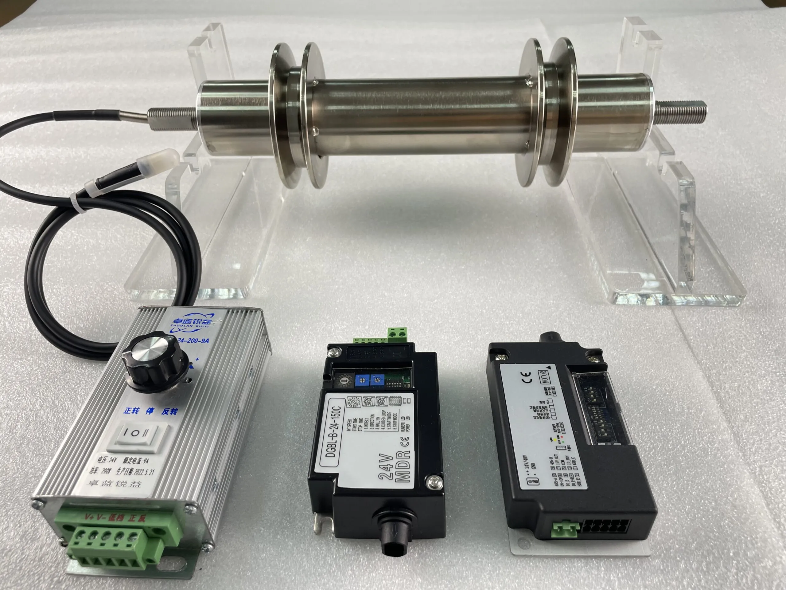 A stainless steel roller with two flanges is on clear stands. Below it are three electronic components: a silver controller with a black knob and Chinese text, and two black modules with green terminals and labels "DGBL-B-24-150C" and "MDR CE".