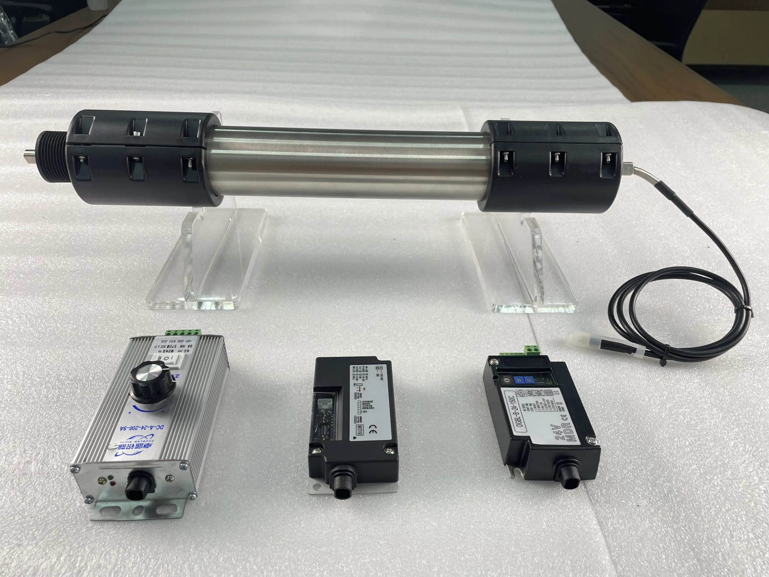 A stainless steel roller with black end caps rests on clear stands. Below it are three electronic control units. The left unit is labeled "DC-A-24-200-9A" and "ZHUOLAN RUIYI". The middle unit is labeled "DGBL-B-24-150C" and "24V MDR CE". The right unit is labeled "DDR-P-100" and "24V MDR CE".