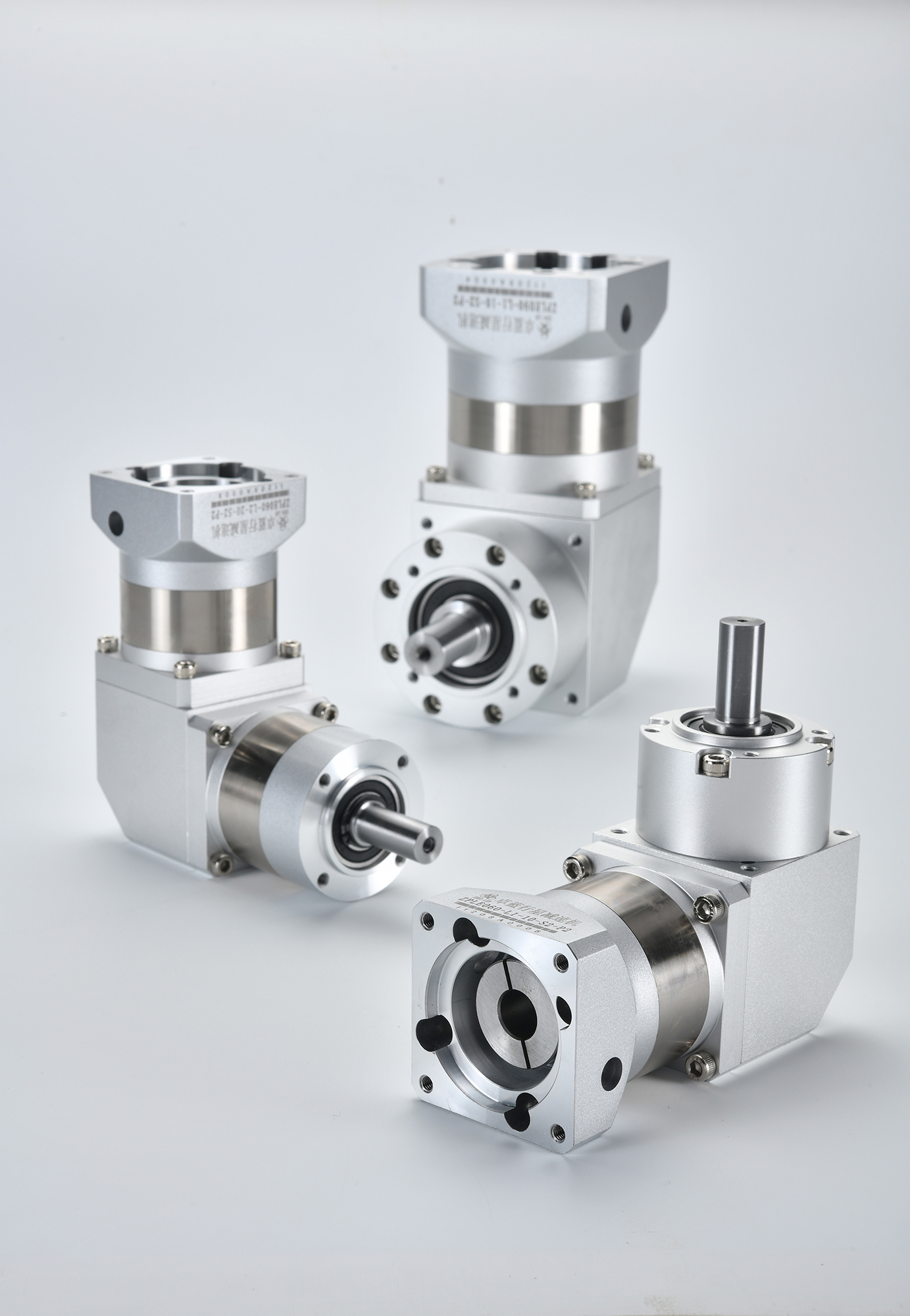 Three silver and gray right-angle planetary gearboxes on a white surface, two with horizontal output shafts and one with a vertical output shaft.