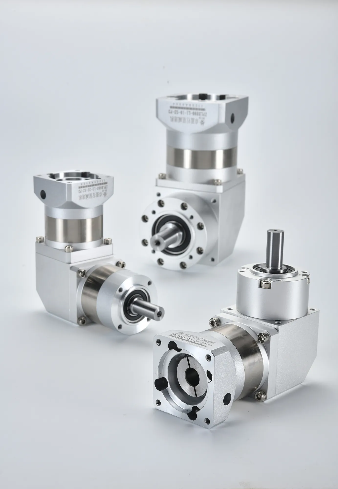 Three silver and gray right-angle planetary gearboxes on a white surface, two with horizontal output shafts and one with a vertical output shaft.
