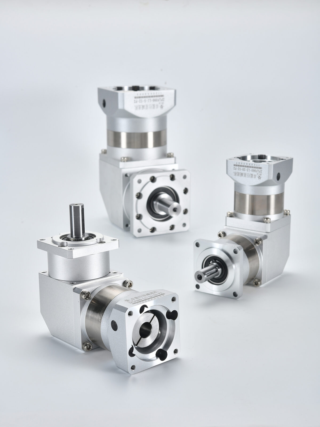 Three silver and gray right-angle planetary gearboxes on a white surface. The front gearbox has a vertical output shaft, while the two in the background have horizontal output shafts. Labels include "ZPLF060-L2-20-S2-P2" and "ZPLF090-L1-5-S2-P2".