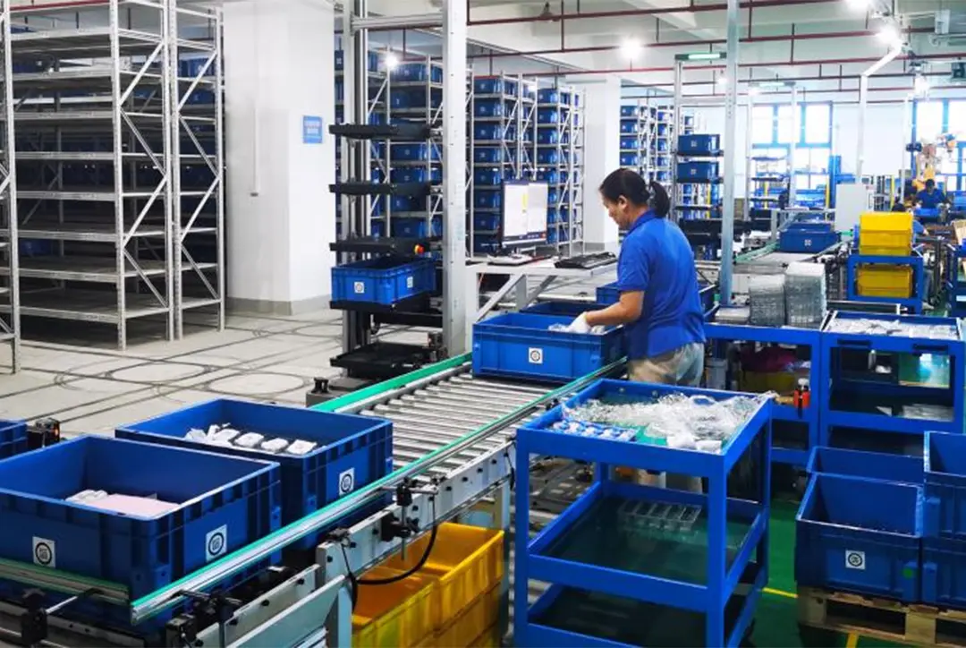 A worker processes blue plastic bins containing components on a conveyor belt system within a warehouse.