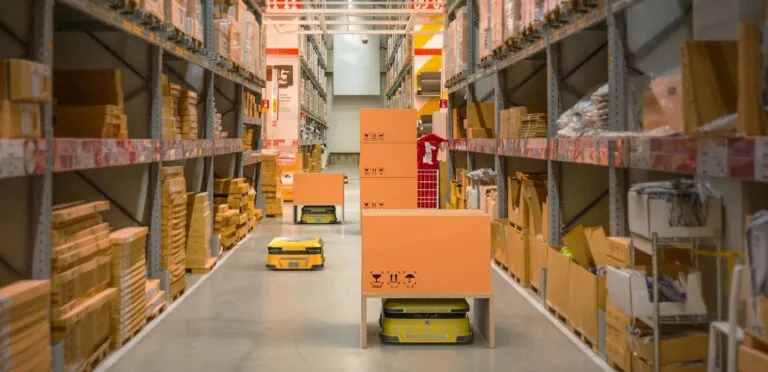 Automated yellow robots transport orange boxes through a warehouse aisle lined with tall storage racks.