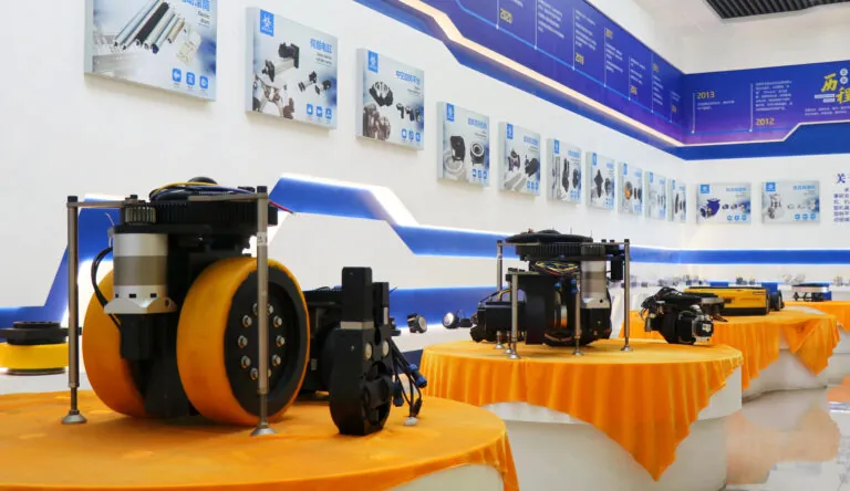 A showroom display featuring several robotic wheel drive units and AGV components arranged on orange-covered tables against a wall with technical product posters.