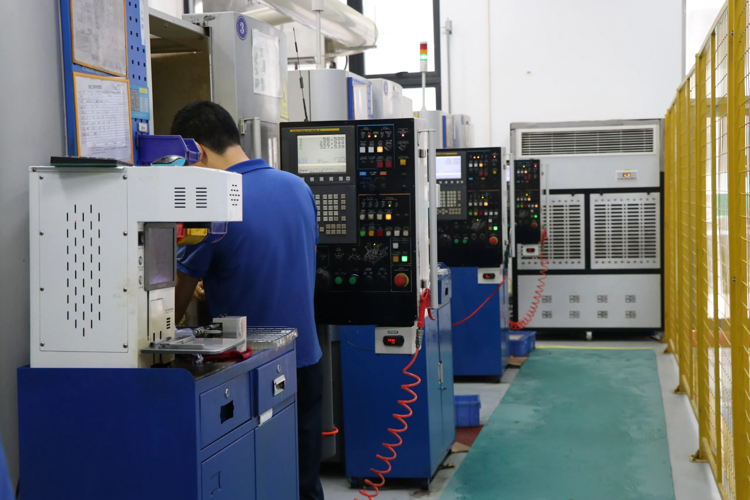 A worker in a blue shirt operating a CNC machine in an industrial workshop with blue cabinets and yellow fencing.
