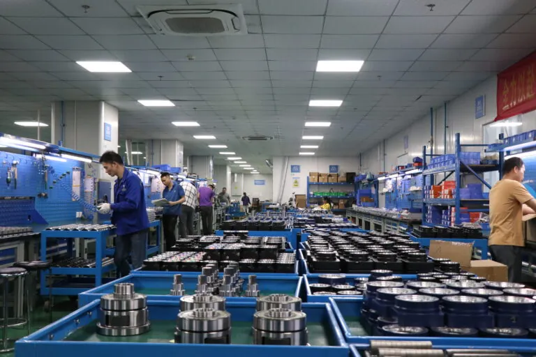 Wide shot of a well-lit factory assembly line with workers in blue uniforms assembling precision metal components.