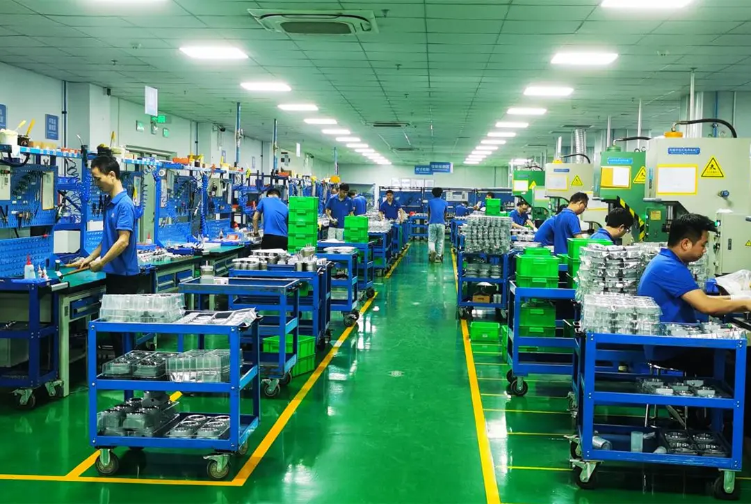 Workers in blue uniforms operate at individual stations along a brightly lit, organized manufacturing assembly line.