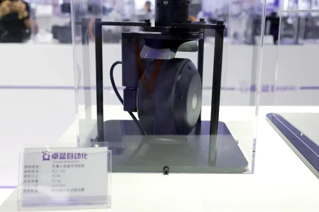 Một bộ phận lái AGV nhỏ gọn với động cơ gắn theo chiều dọc được đặt bên trong hộp trưng bày trong suốt trên bàn màu trắng.