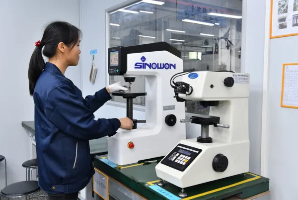A woman in a blue lab coat operating a Sinowon hardness testing machine in a factory setting, with another testing machine nearby. The screen displays a reading of 57.0.