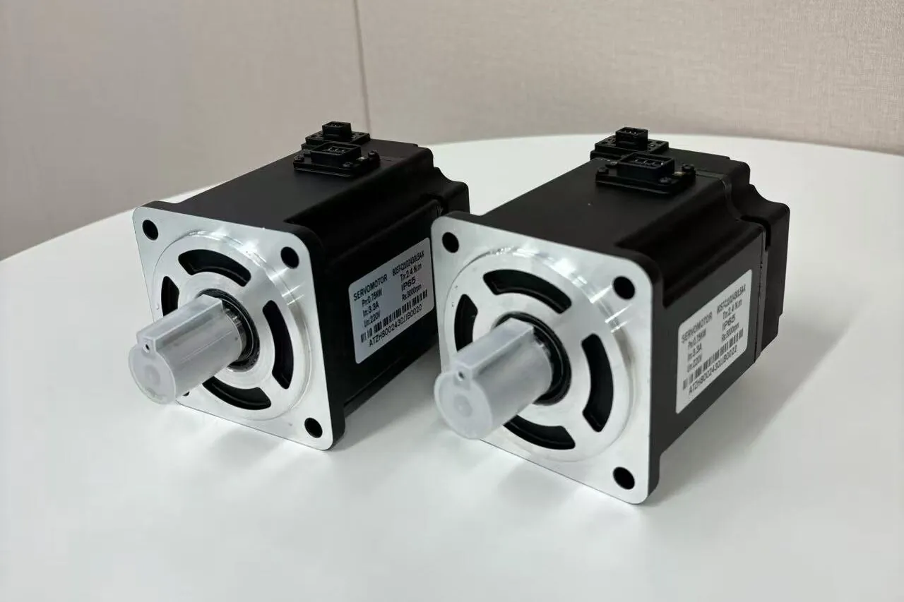 Two black servomotors with silver faces and shafts are displayed on a white table. Each motor has a label with "SERVOMOTOR" and specifications.