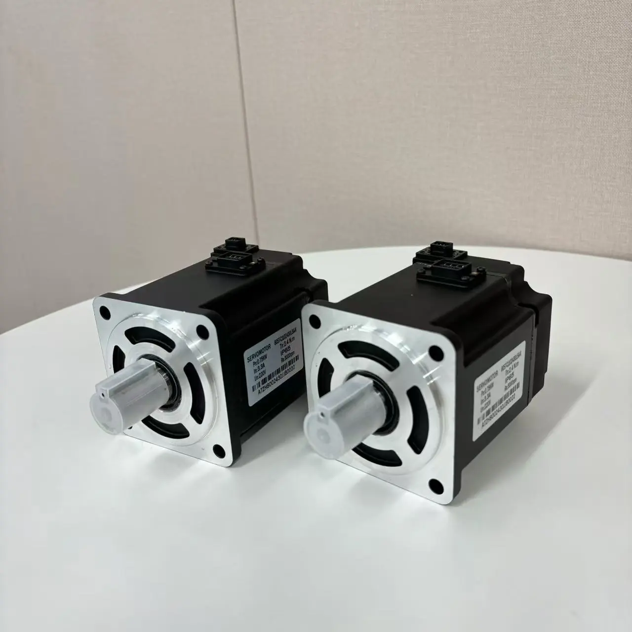 Two black servomotors with silver faces and shafts are displayed on a white table. Each motor has a label with "SERVOMOTOR" and specifications.