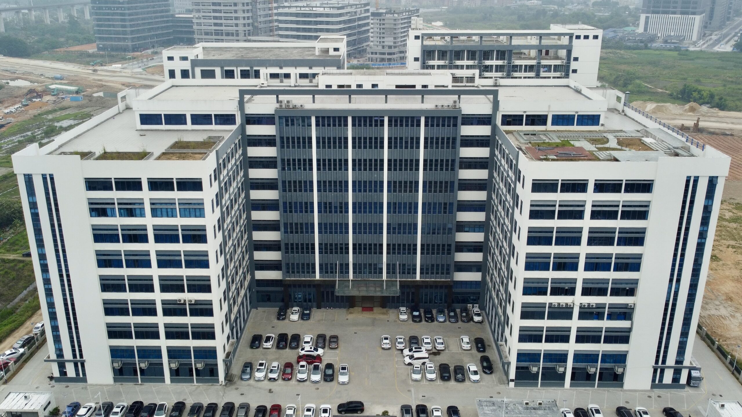 An aerial view of a large, modern white and gray building with blue-tinted windows, surrounded by a parking lot full of cars. Construction sites and other buildings are visible in the background.
