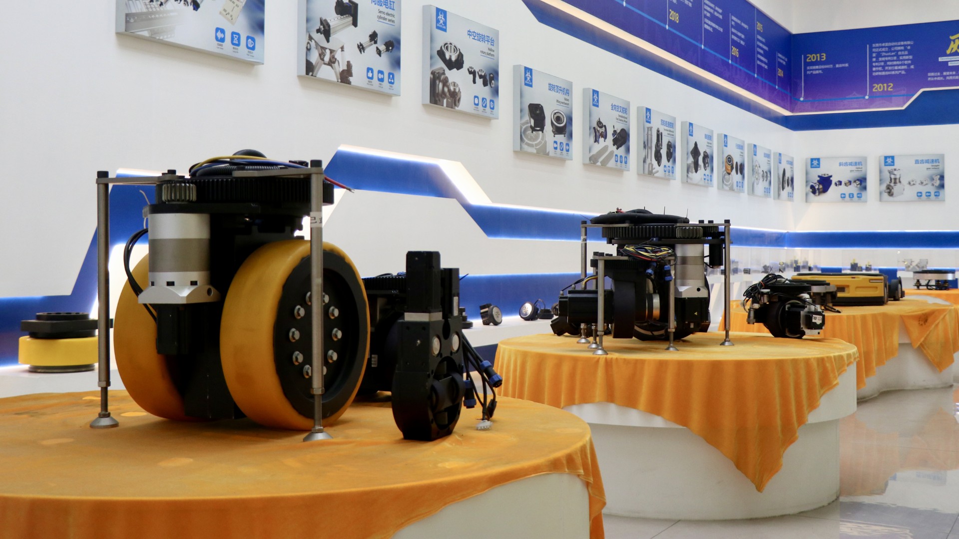 Industrial robotic drive wheels on orange cloth-covered pedestals in a technology exhibition showroom.
