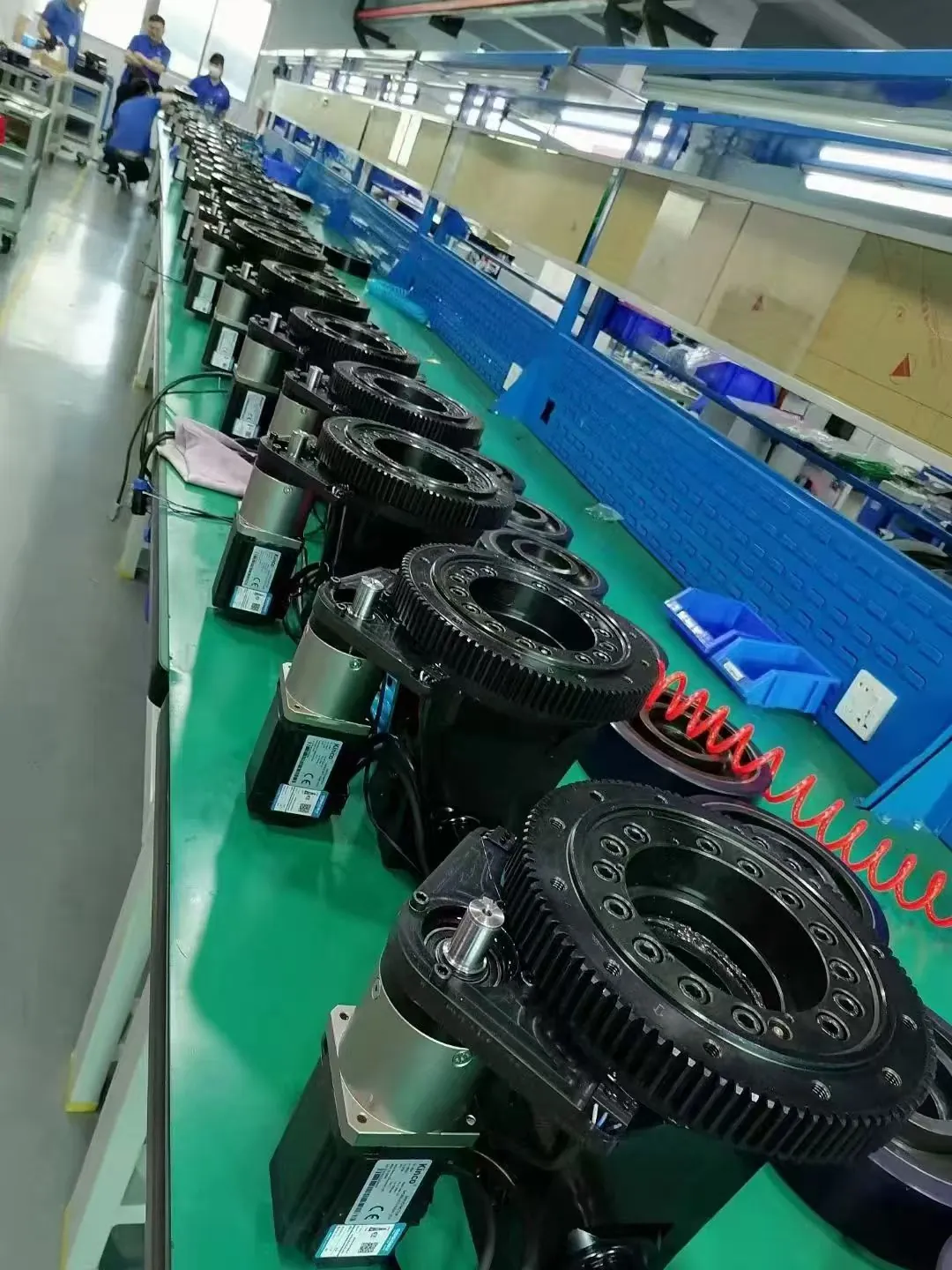 Một dây chuyền lắp ráp gồm nhiều cụm bánh dẫn động AGV (có các vòng bánh răng màu đen cỡ lớn và động cơ) được bố trí trên một chiếc bàn làm việc dài màu xanh lá cây bên trong nhà máy.