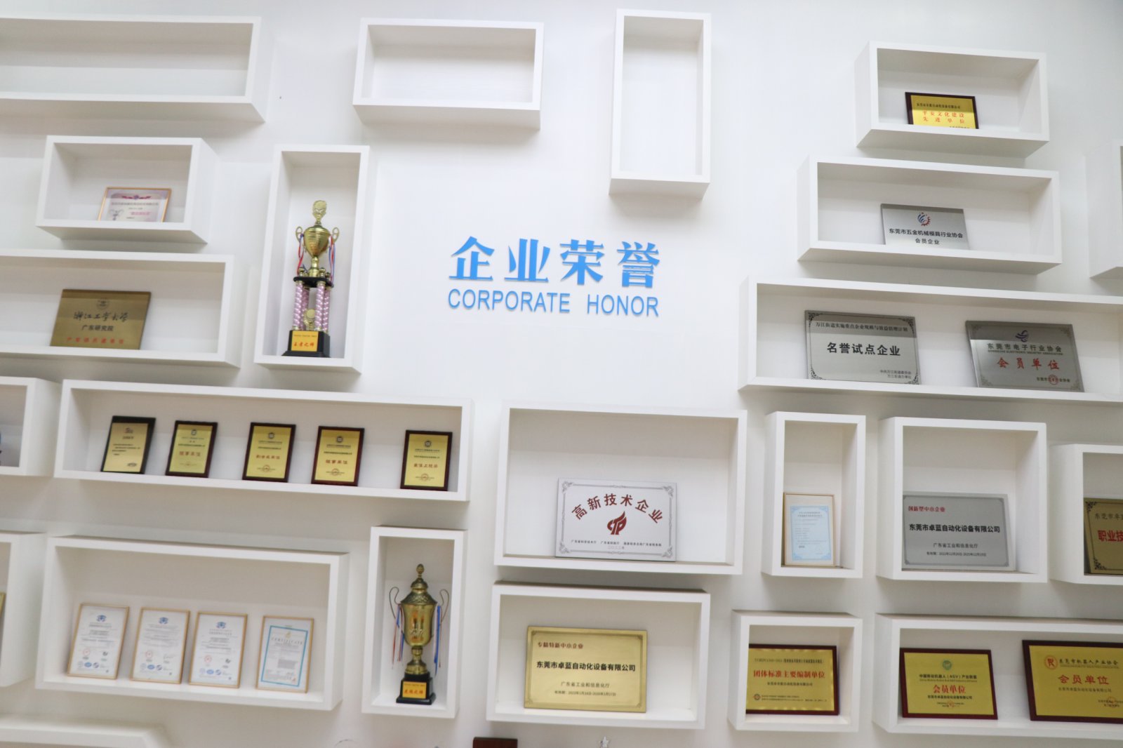 A white wall with "CORPORATE HONOR" in blue text, surrounded by white rectangular shelves displaying trophies and various framed certificates.