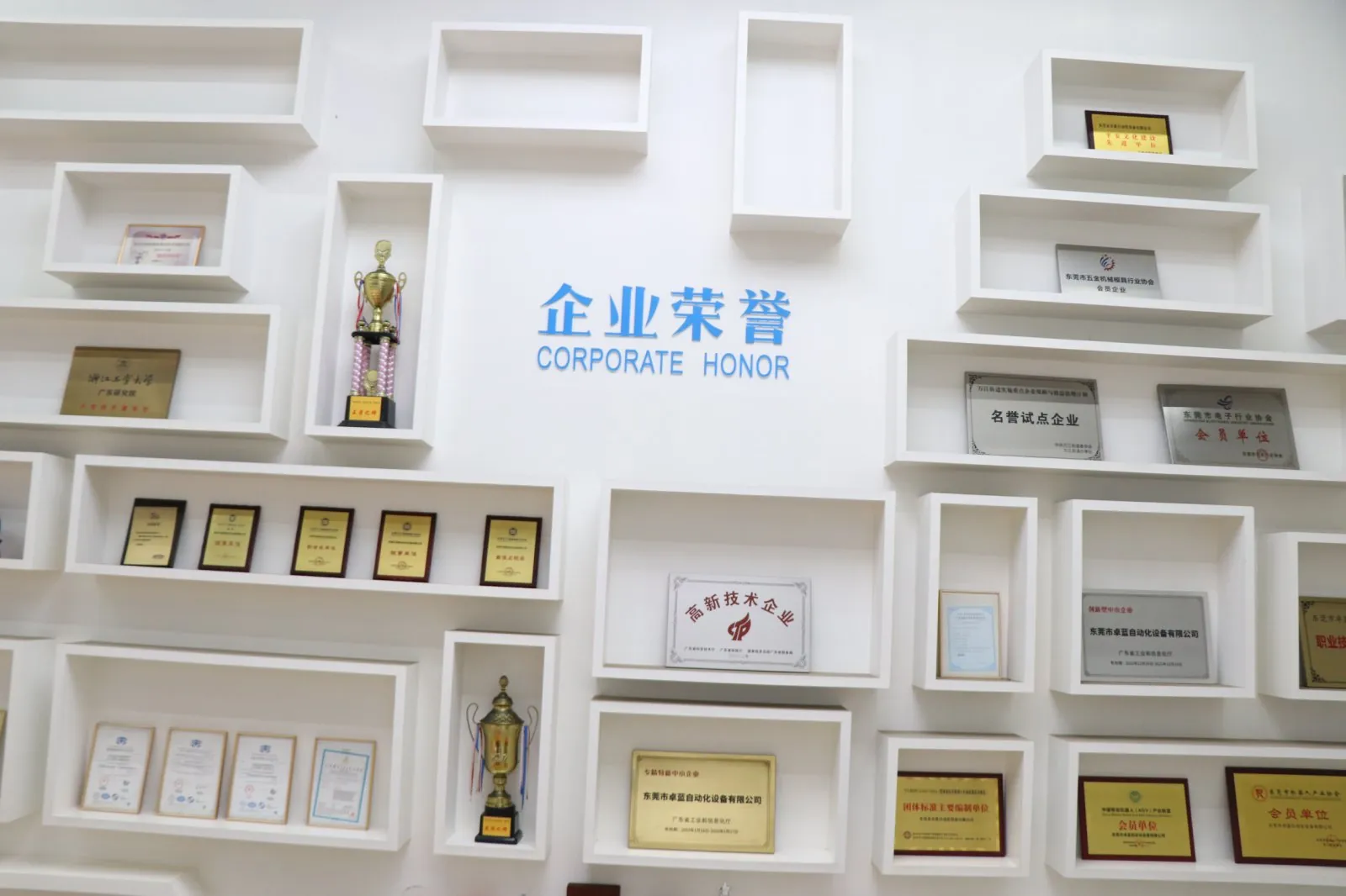 A white wall with "CORPORATE HONOR" in blue text, surrounded by white rectangular shelves displaying trophies and various framed certificates.