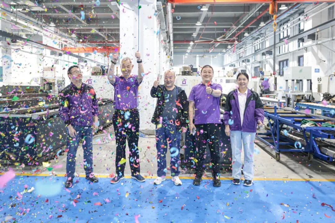 Five people, three men and two women, stand in a factory with colorful confetti falling around them. They are smiling and giving thumbs up.