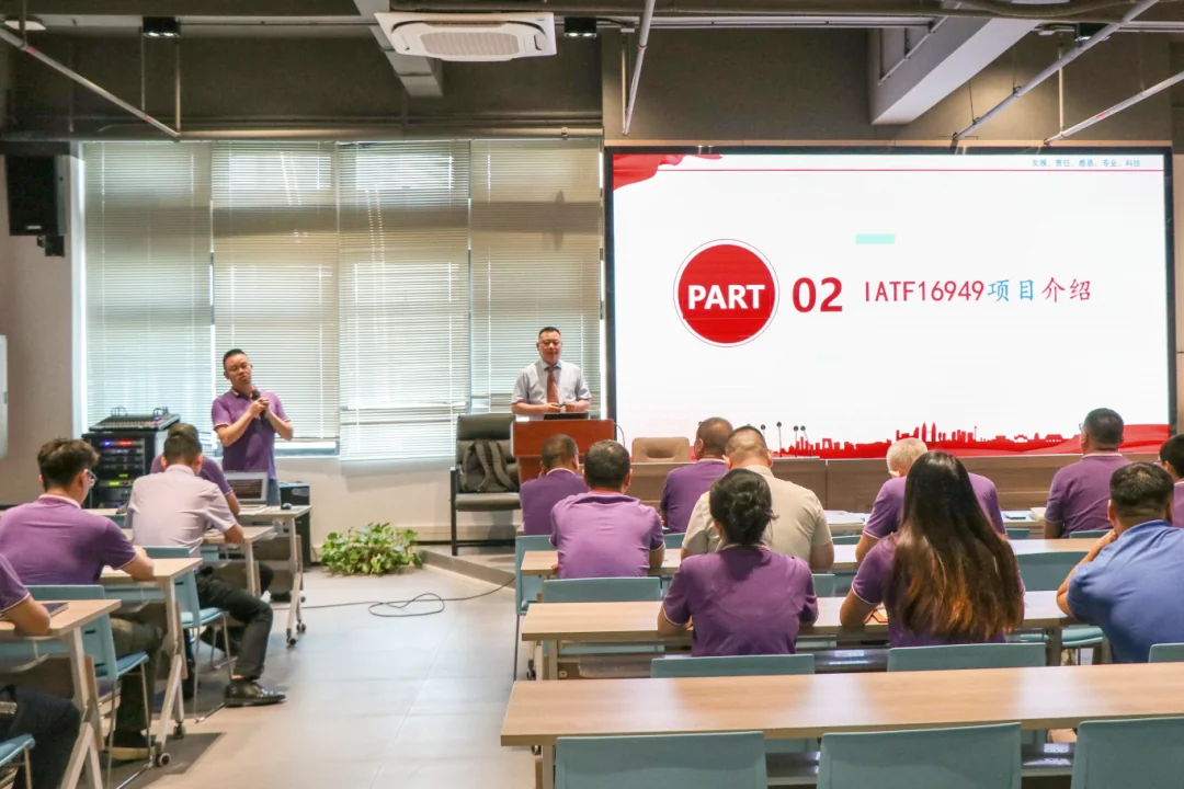 A man in a purple shirt speaks into a microphone, while another man stands at a podium. An audience in purple shirts watches a screen displaying "PART 02 IATF16949".