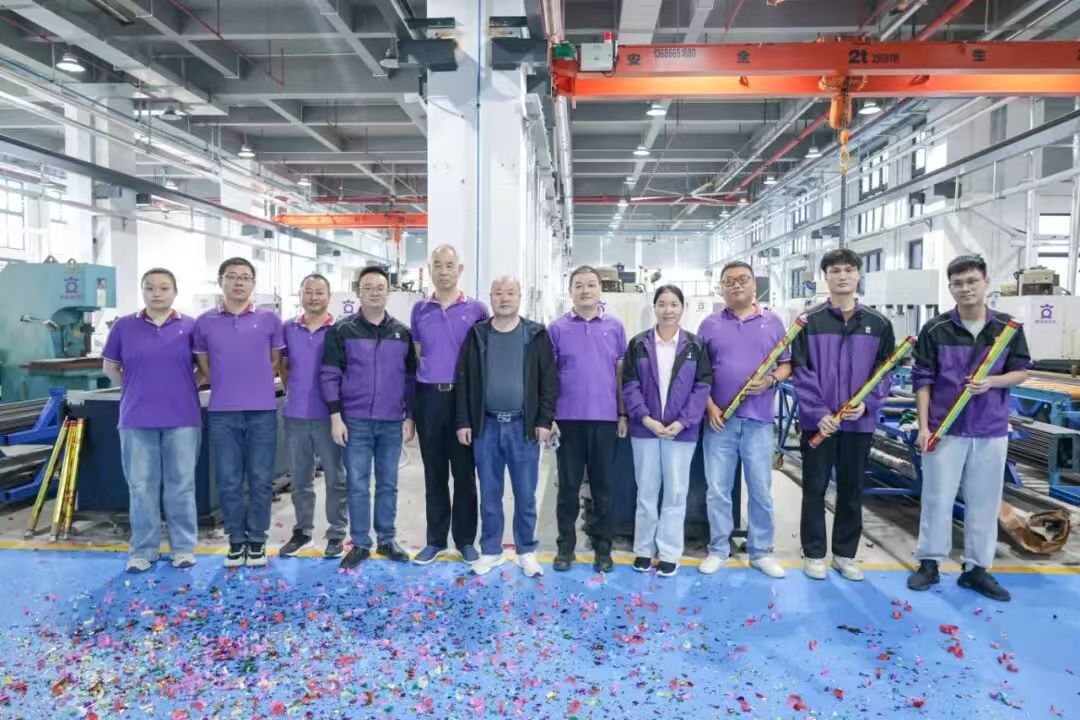 A group of 12 factory workers, mostly men, in purple shirts and jackets, stand in a line on a blue floor covered in confetti. Two men on the right hold yellow and red confetti cannons. An orange overhead crane with "2T" and Chinese characters is visible above.