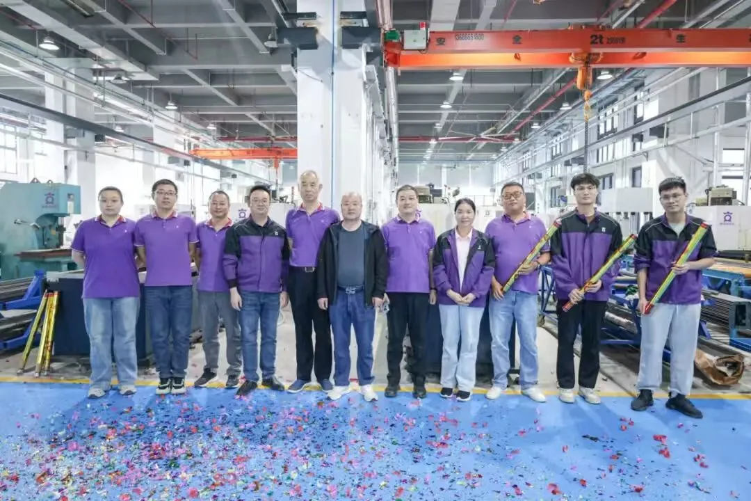 A group of 12 factory workers, mostly men, in purple shirts and jackets, stand in a line on a blue floor covered in confetti. Two men on the right hold yellow and red confetti cannons. An orange overhead crane with "2T" and Chinese characters is visible above.