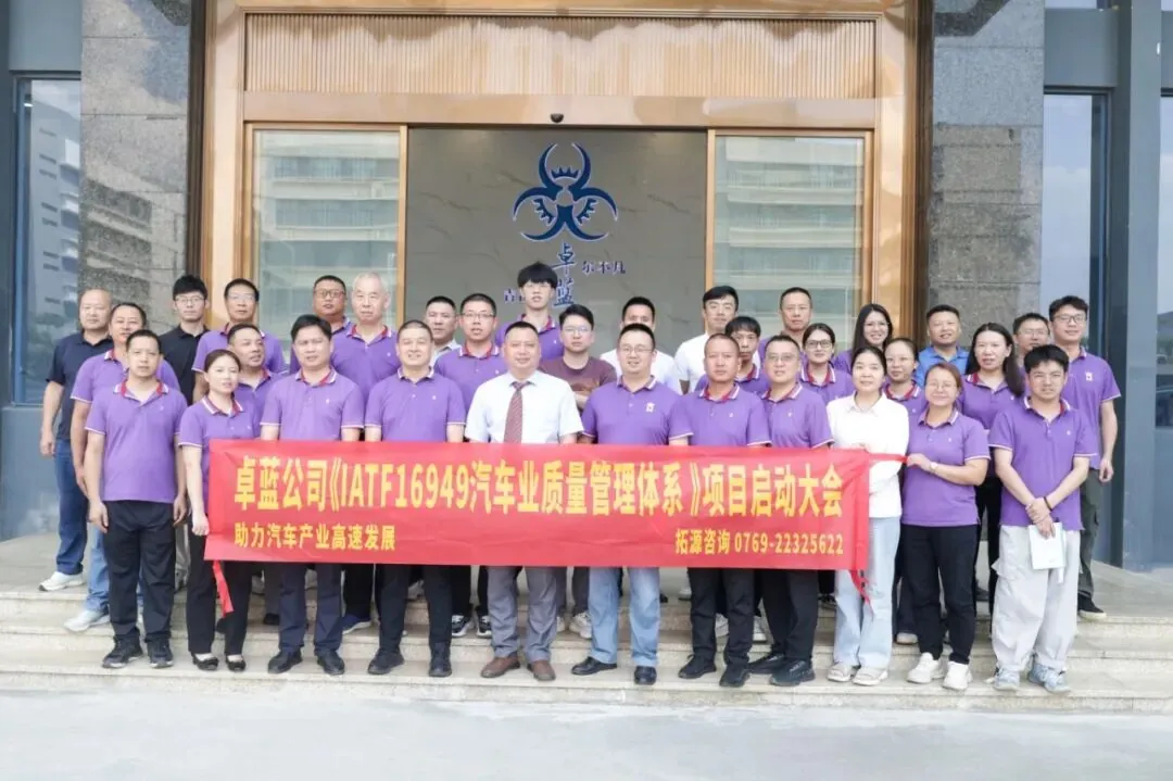 A large group of people, mostly men, wearing purple polo shirts, stand on steps in front of a building. They hold a red banner with yellow text.