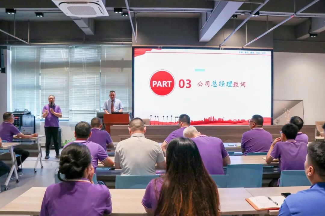 A man in a purple shirt speaks into a microphone, while another man stands at a podium. An audience in purple shirts watches a screen displaying "PART 03".