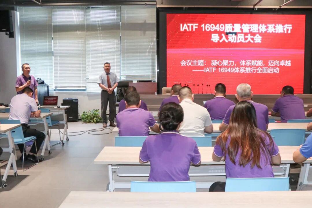 A man in a purple shirt speaks into a microphone, while another man stands at the front. An audience watches a screen displaying "IATF 16949".