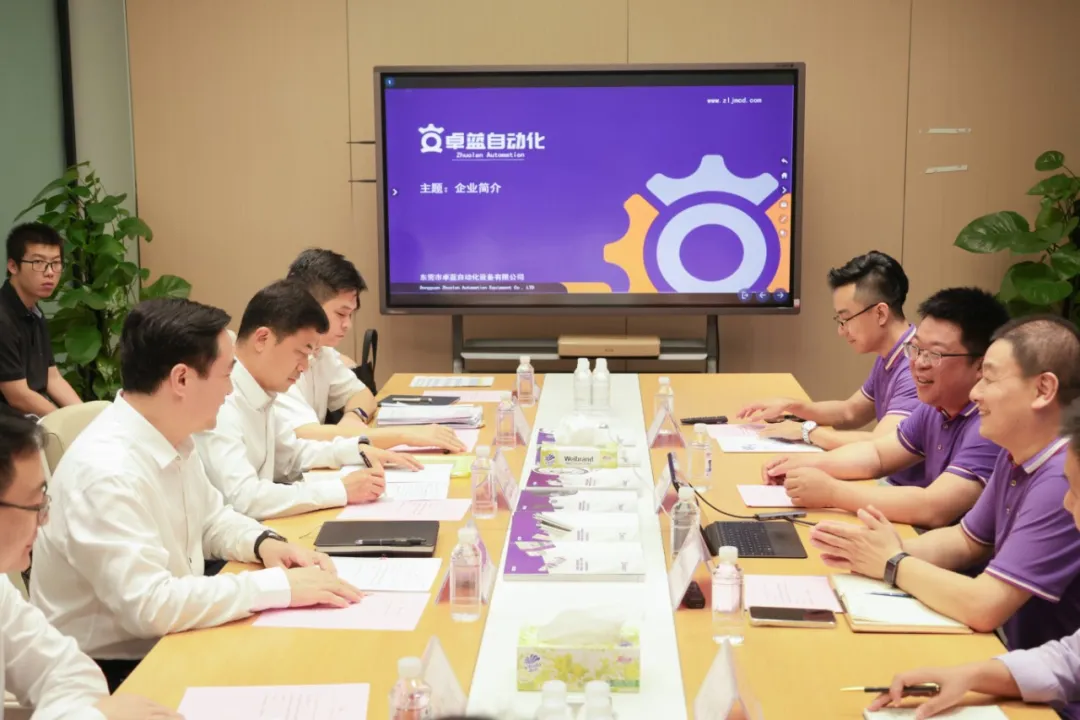 A professional meeting in a conference room with two groups of men seated across a table, facing a large display screen.