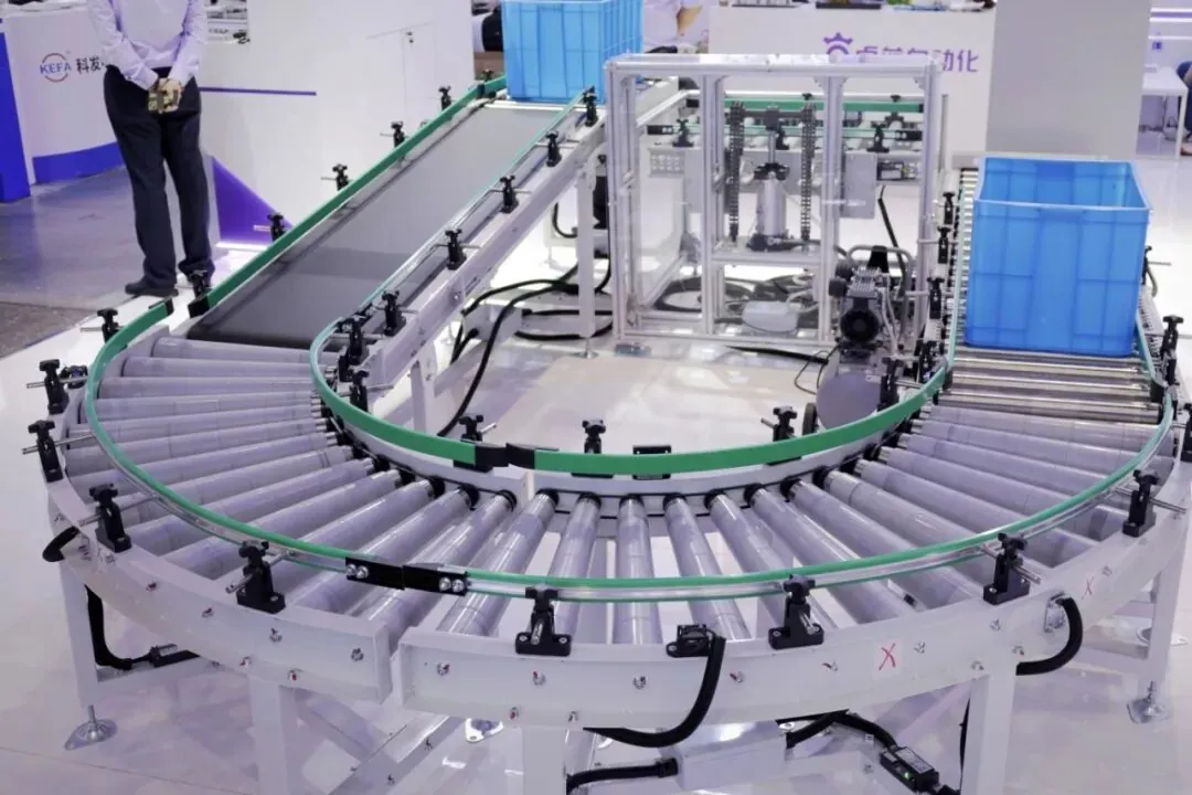 An automated curved roller conveyor system with green side guardrails and blue plastic crates on a trade show floor.