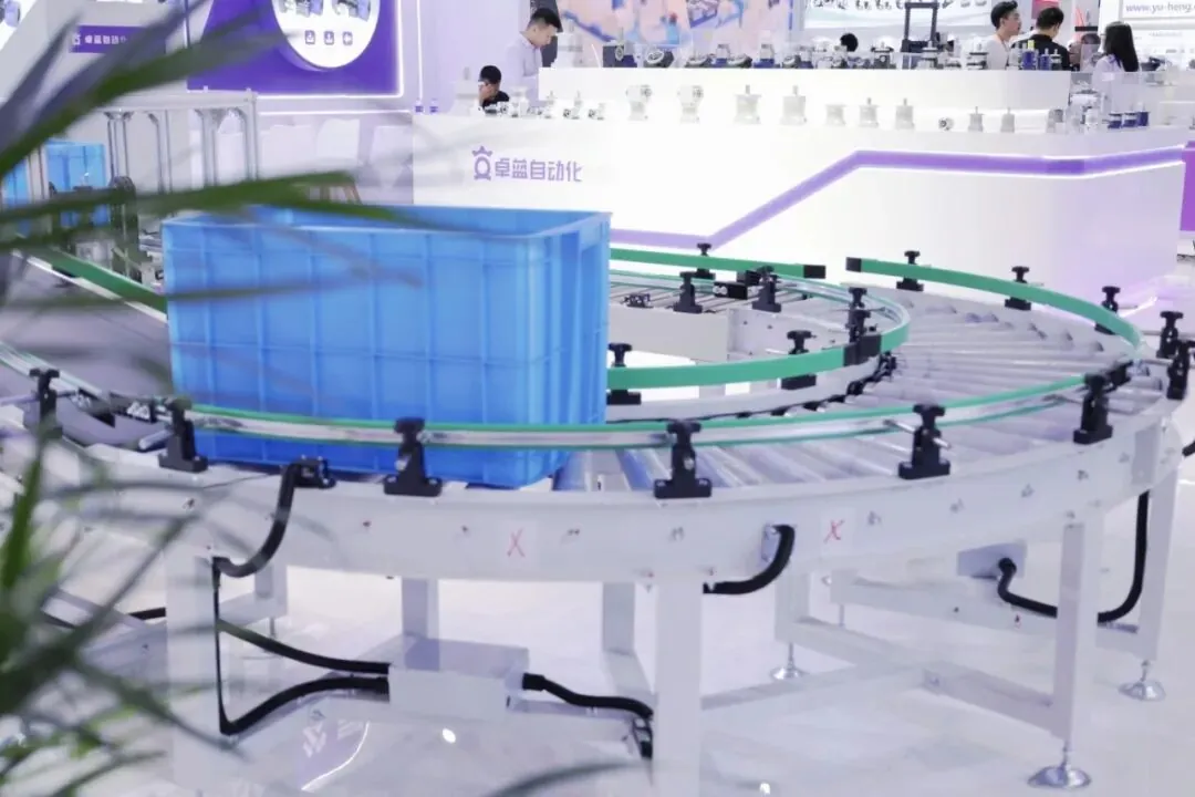 A blue plastic crate moving along a curved motorized roller conveyor track at a trade show exhibition.